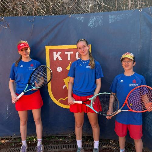 3 Kinder mit Tennisschläger vor Tennisplatzbegrenzung